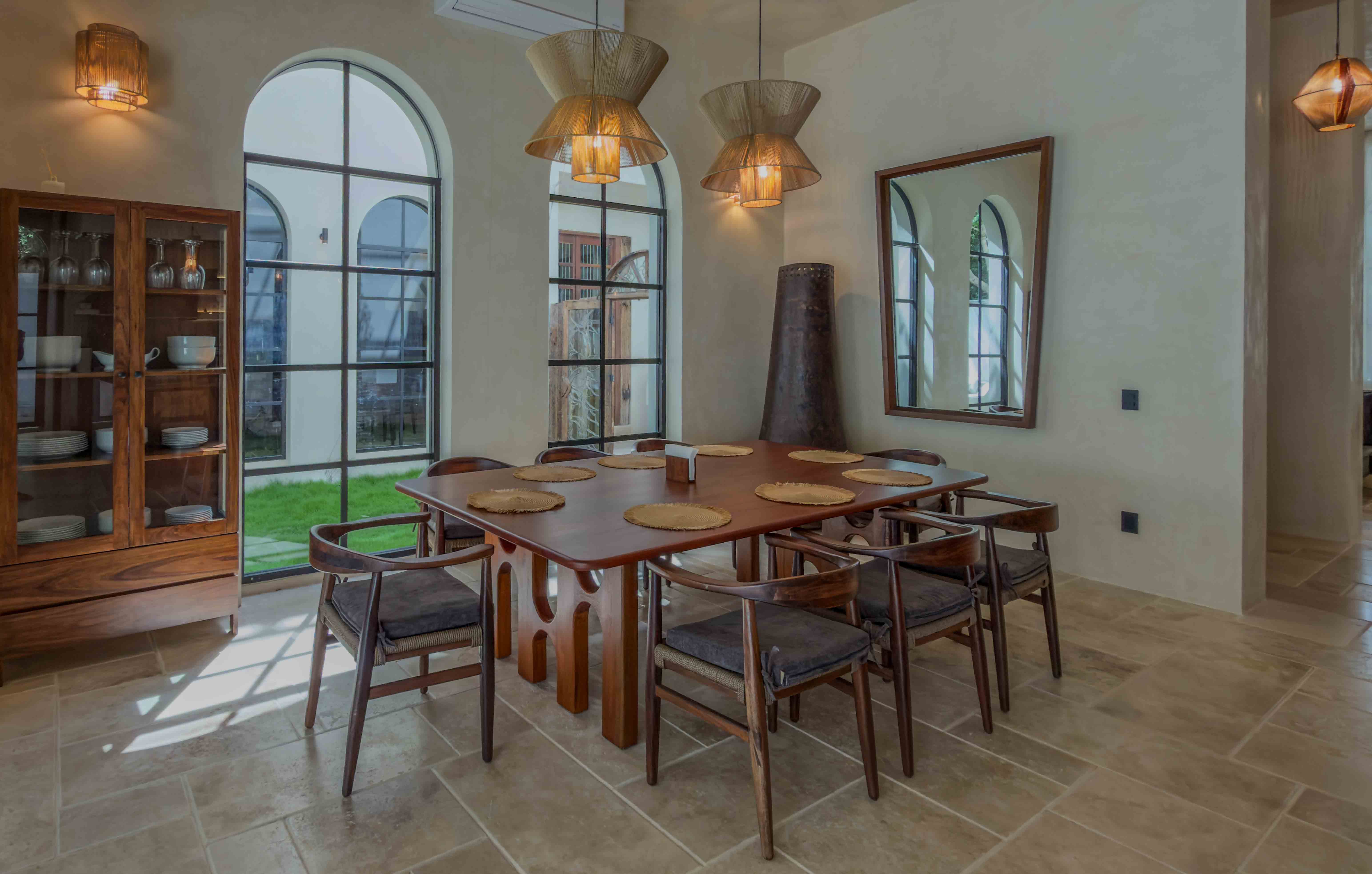 Large wooden dining table with designer chairs and arched windows in a sunlit interior.