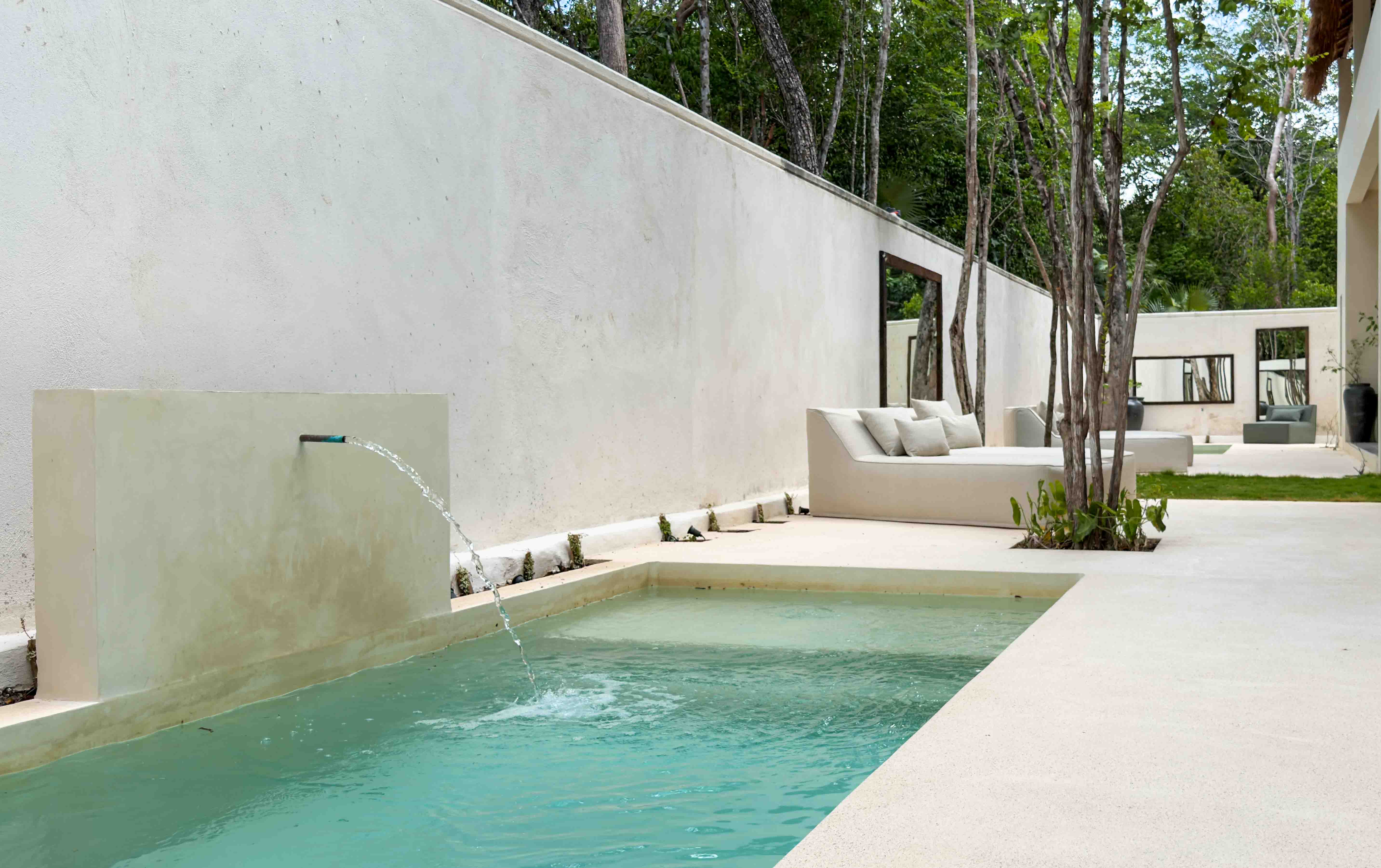 Minimalist lap pool with water feature and lounge seating against a white limestone wall.