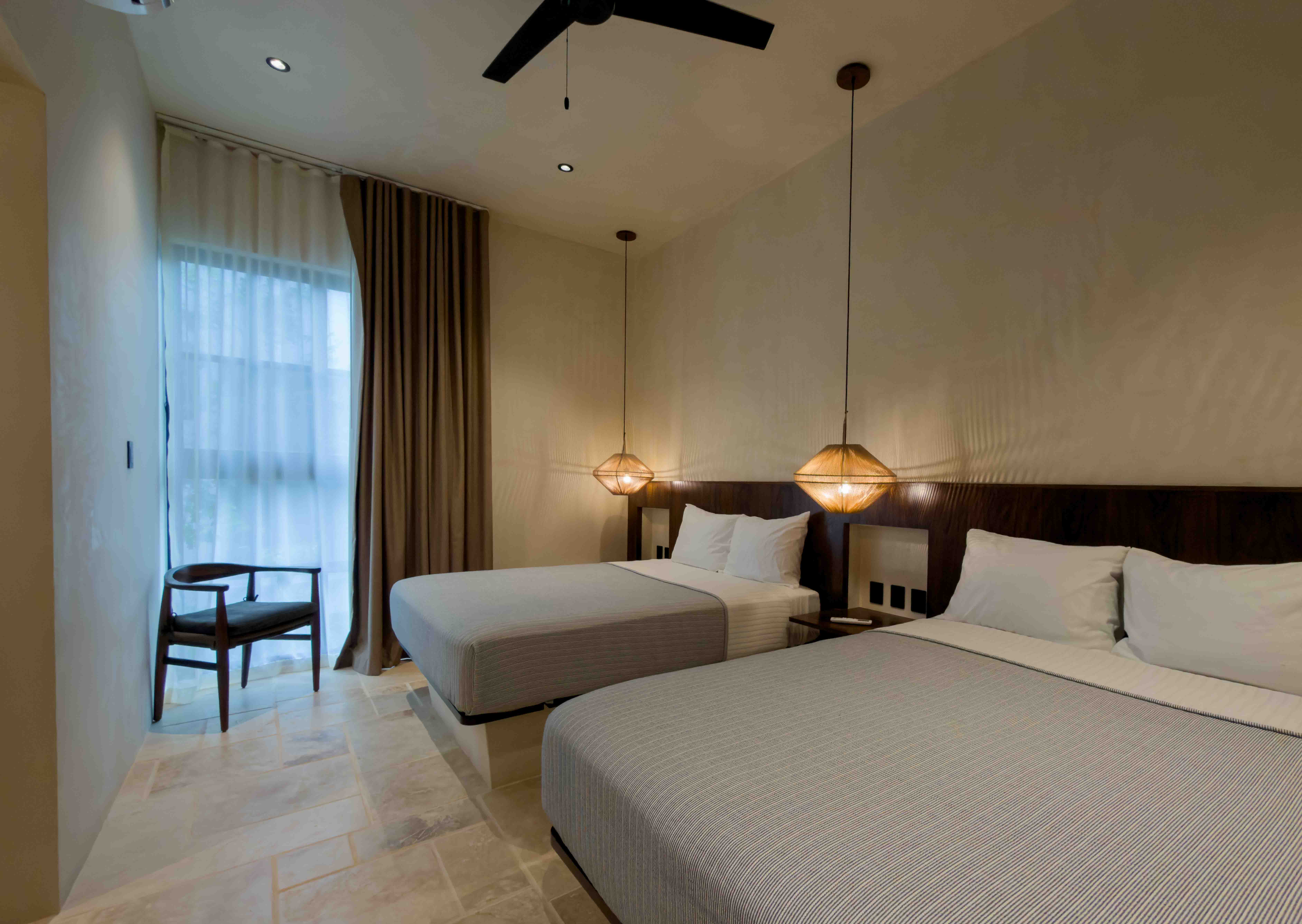 Minimalist suite with two queen beds, warm pendant lighting, and neutral stone flooring.